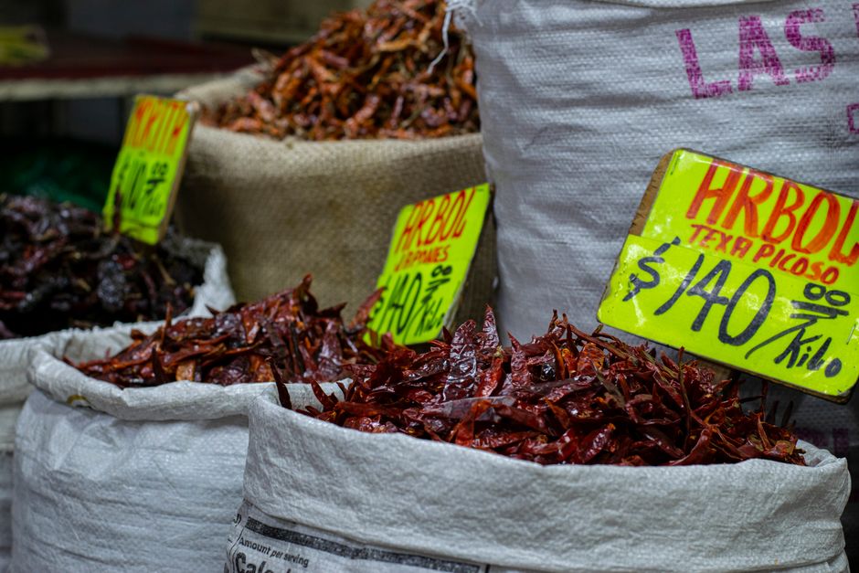 Close-up de pimentas secas em sacos num mercado, com etiquetas de preço escritas à mão em cores vibrantes.